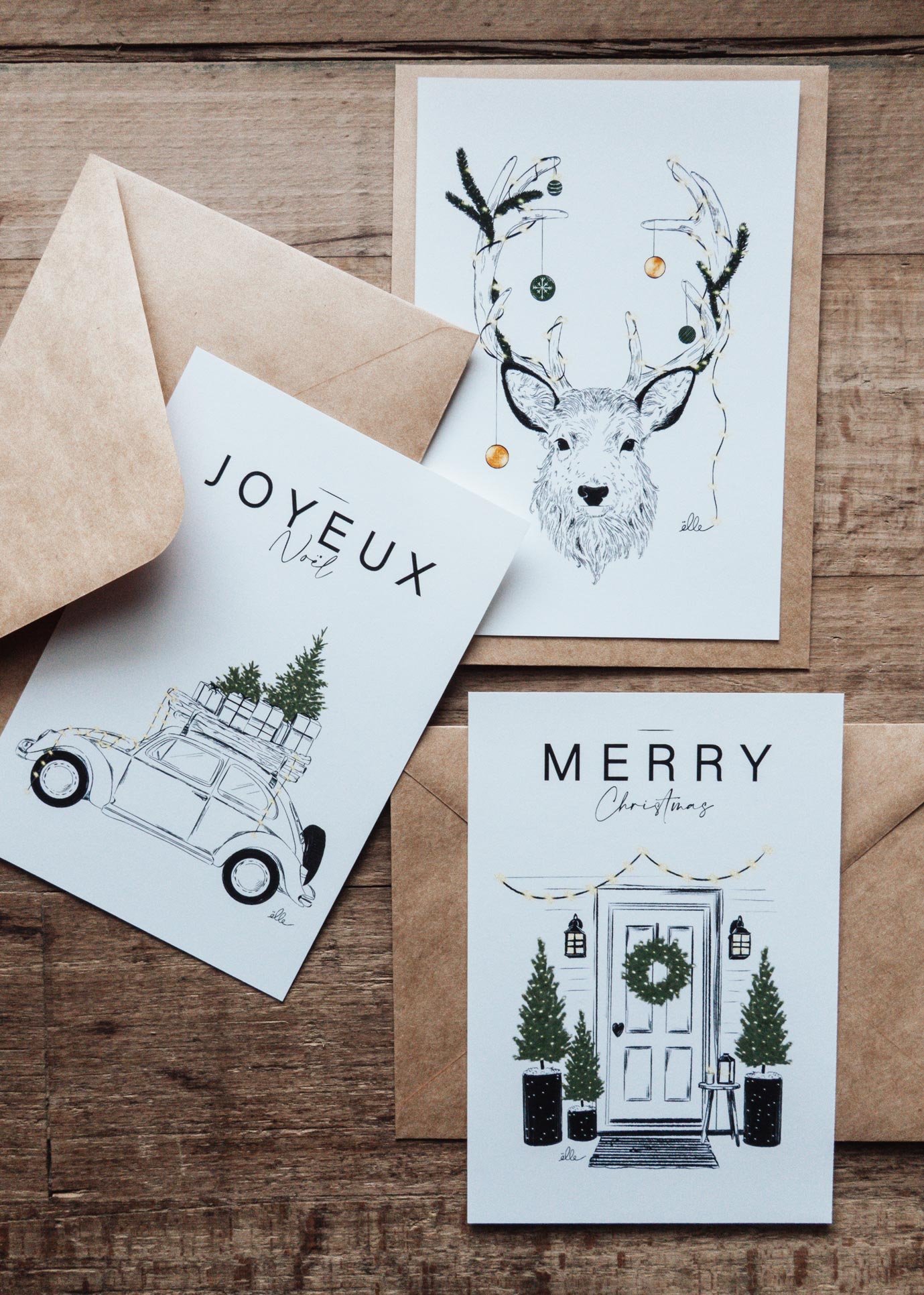 Cartes de vœux de Noël illustrées, posées sur une table en bois, comprenant une carte “Joyeux Noël” avec une voiture chargée de cadeaux, une carte “Merry Christmas” avec une porte décorée, et une carte avec un cerf aux bois ornés, accompagnées d’enveloppes kraft.