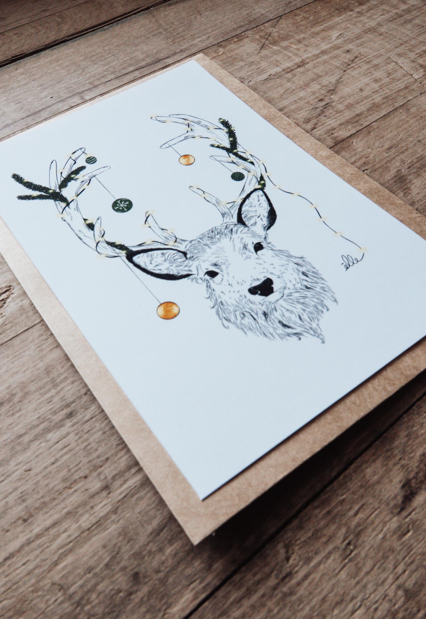 Cartes de vœux de Noël illustrées, posées sur une table en bois, comprenant une carte “Joyeux Noël” avec une voiture chargée de cadeaux, une carte “Merry Christmas” avec une porte décorée, et une carte avec un cerf aux bois ornés, accompagnées d’enveloppes kraft.