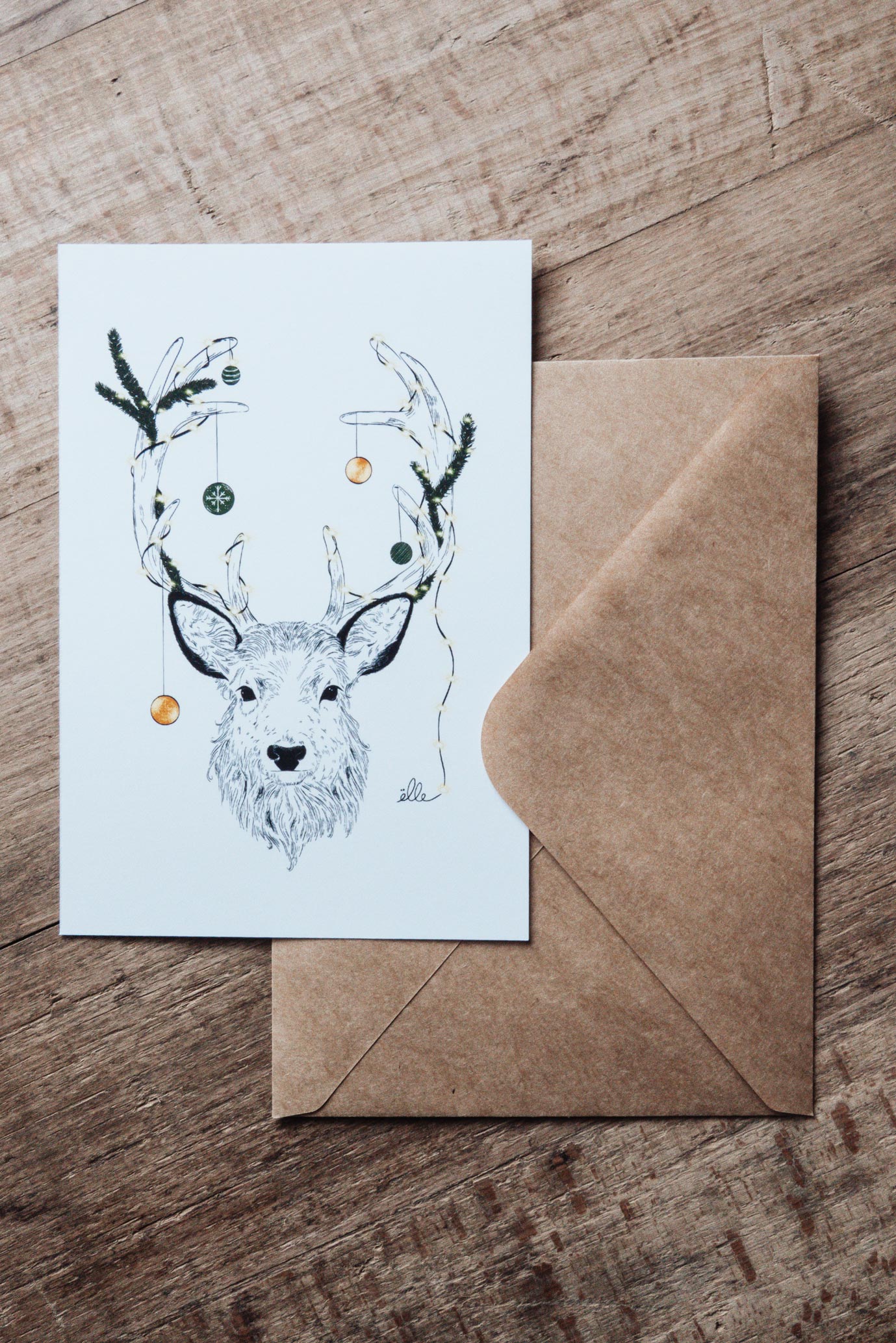 Cartes de vœux de Noël illustrées, posées sur une table en bois, comprenant une carte “Joyeux Noël” avec une voiture chargée de cadeaux, une carte “Merry Christmas” avec une porte décorée, et une carte avec un cerf aux bois ornés, accompagnées d’enveloppes kraft.