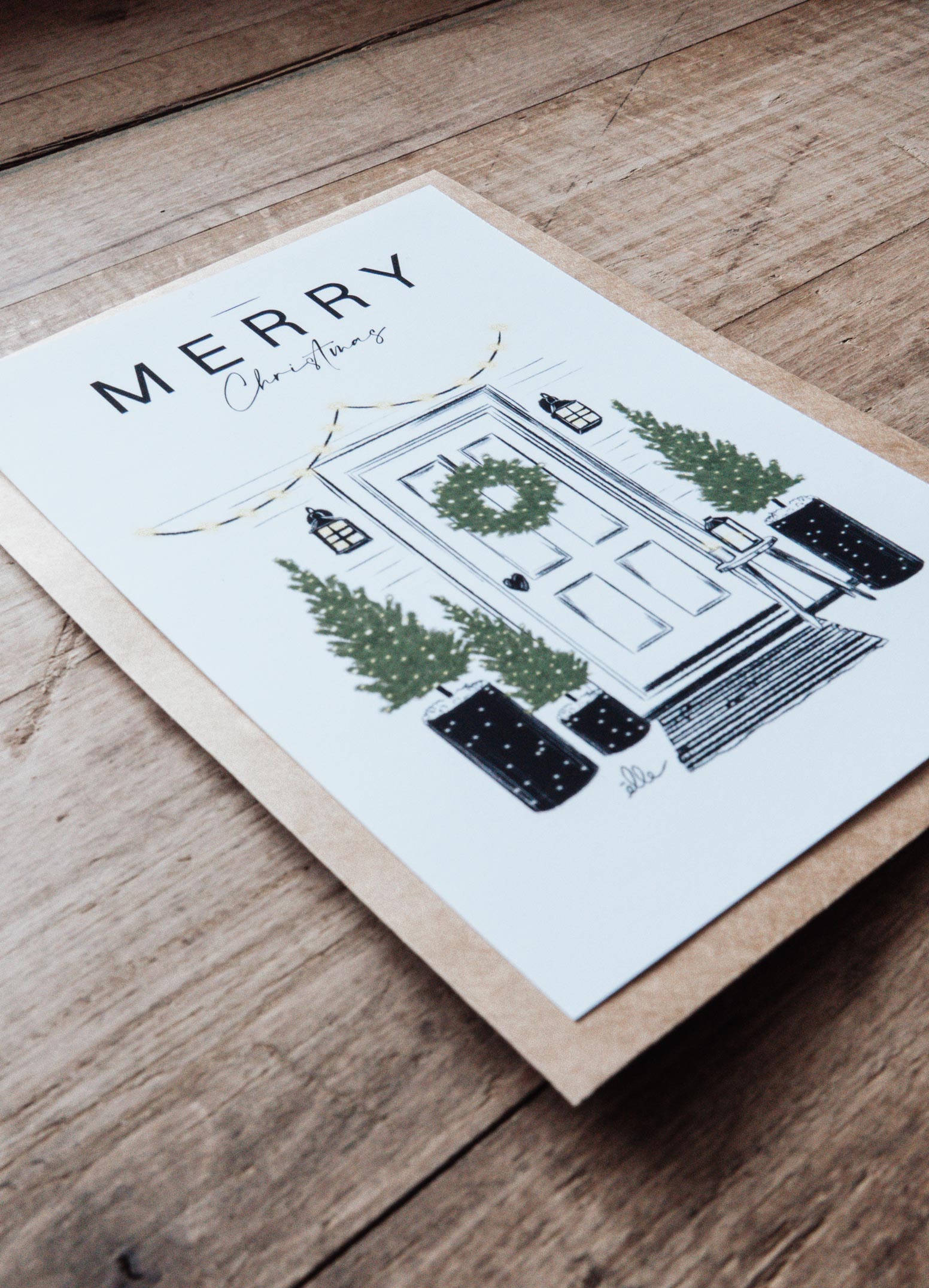 Cartes de vœux de Noël illustrées, posées sur une table en bois, comprenant une carte “Joyeux Noël” avec une voiture chargée de cadeaux, une carte “Merry Christmas” avec une porte décorée, et une carte avec un cerf aux bois ornés, accompagnées d’enveloppes kraft.