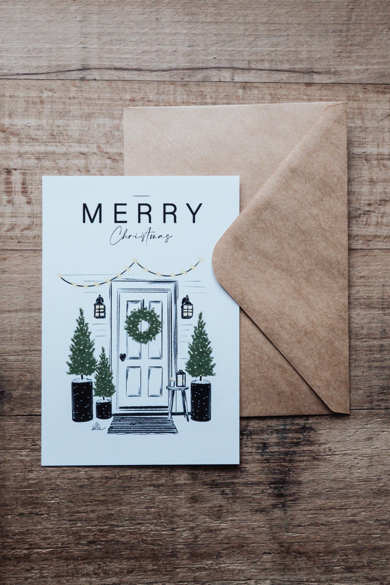 Cartes de vœux de Noël illustrées, posées sur une table en bois, comprenant une carte “Joyeux Noël” avec une voiture chargée de cadeaux, une carte “Merry Christmas” avec une porte décorée, et une carte avec un cerf aux bois ornés, accompagnées d’enveloppes kraft.