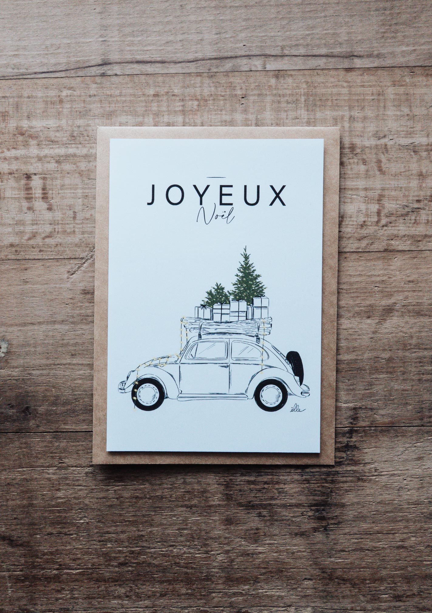 Cartes de vœux de Noël illustrées, posées sur une table en bois, comprenant une carte “Joyeux Noël” avec une voiture chargée de cadeaux, une carte “Merry Christmas” avec une porte décorée, et une carte avec un cerf aux bois ornés, accompagnées d’enveloppes kraft.