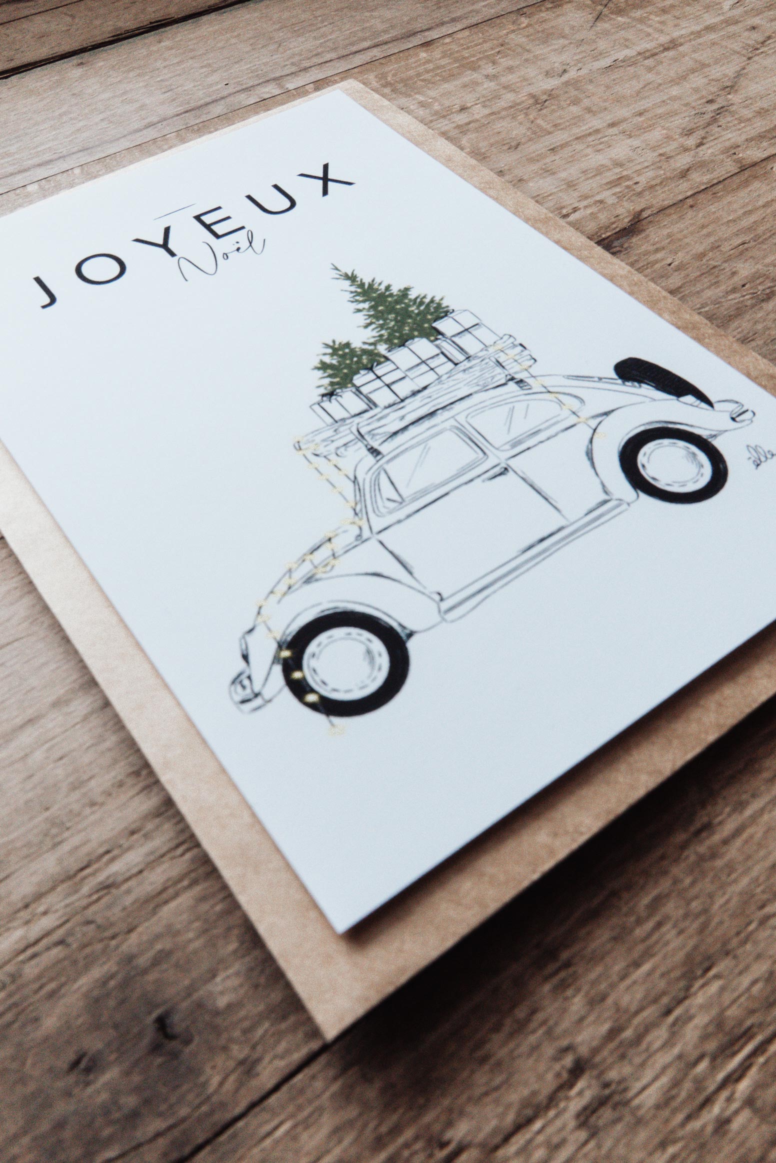 Cartes de vœux de Noël illustrées, posées sur une table en bois, comprenant une carte “Joyeux Noël” avec une voiture chargée de cadeaux, une carte “Merry Christmas” avec une porte décorée, et une carte avec un cerf aux bois ornés, accompagnées d’enveloppes kraft.