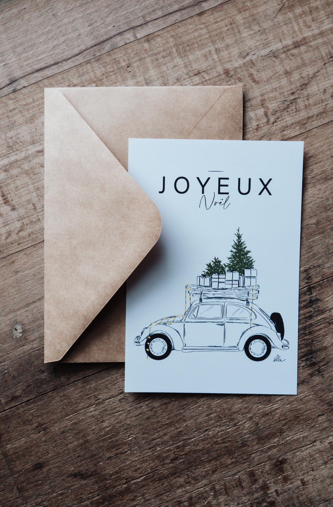 Cartes de vœux de Noël illustrées, posées sur une table en bois, comprenant une carte “Joyeux Noël” avec une voiture chargée de cadeaux, une carte “Merry Christmas” avec une porte décorée, et une carte avec un cerf aux bois ornés, accompagnées d’enveloppes kraft.